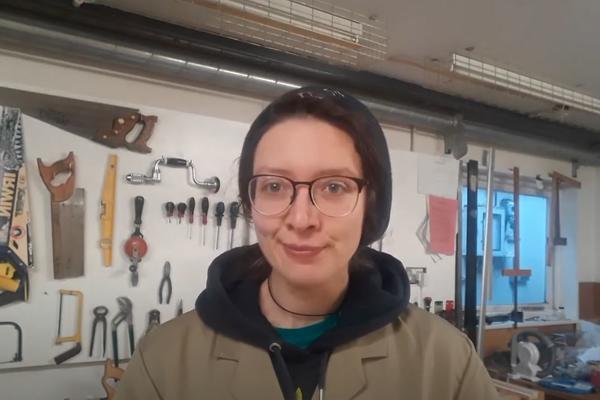 A woman speaks to a webcam from a technician's workshop