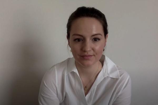 A young woman speaks to a webcam, with a white background