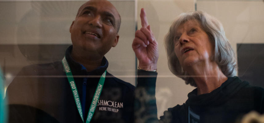 Museum staff member and volunteer stand and look through a glass case of museum objects